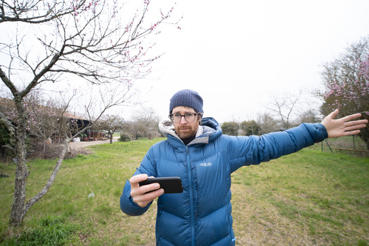 Man in blue winter jacket holding smartphone while gesturing outdoors in open field.