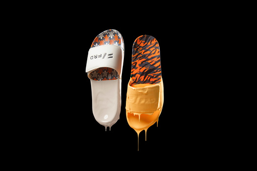 Two white slide sandals with orange tiger-stripe pattern dripping onto cream-colored soles against black background.