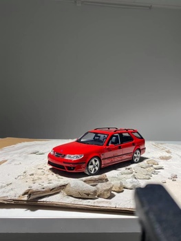 Red station wagon model car positioned on a map with stones, casting a shadow under studio lighting.