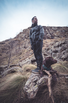 Hiker standing on a rocky outcrop against a barren hillside under overcast sky.