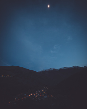 Moonlit mountain landscape with silhouetted peaks and scattered lights from a valley settlement below.