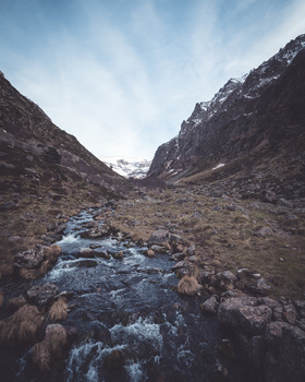 Alpine stream flowing through a rocky valley between steep mountain slopes with snow at higher elevations.