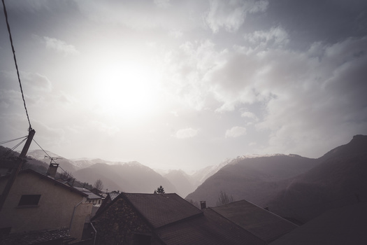Alpine valley shrouded in atmospheric haze with sun breaking through clouds above mountain ridges.