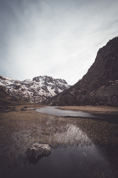 Snow-capped mountains frame a still water body reflecting gray sky in a barren valley.