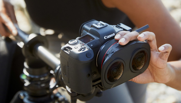 Hands holding a Canon cinema camera with dual lens mounts, with a tripod-mounted cinema lens visible in the background.