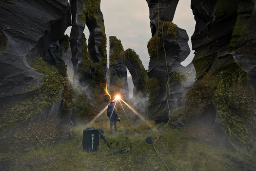 Photographer standing in misty gorge surrounded by moss-covered rock formations, illuminated by golden light from above.