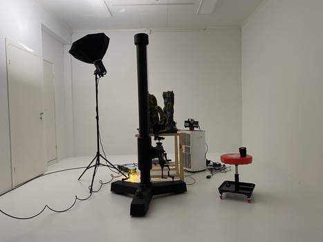 Professional studio setup with tripod-mounted softbox, articulating arm, mechanical equipment on platform, and red rolling stool in white room.
