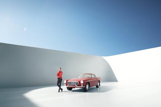 Person in red jacket standing beside a red vintage car against curved white architectural walls under clear blue sky.