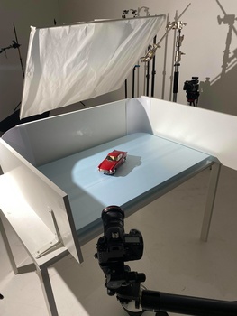 Product photography setup with a red toy car on a light blue surface inside a white shooting tent, with camera on tripod and diffusion panel visible.