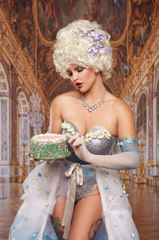 Woman in elaborate costume with ornate white headpiece and jeweled accessories poses against baroque architectural backdrop.