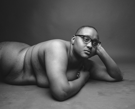 Shirtless man wearing glasses in prone position, resting on forearms against neutral background.