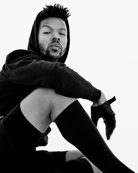 Man in black hoodie posed against white background, looking over shoulder at camera.
