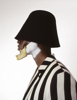 Profile view of person wearing oversized black hat and striped shirt with geometric gold and white face sculpture.