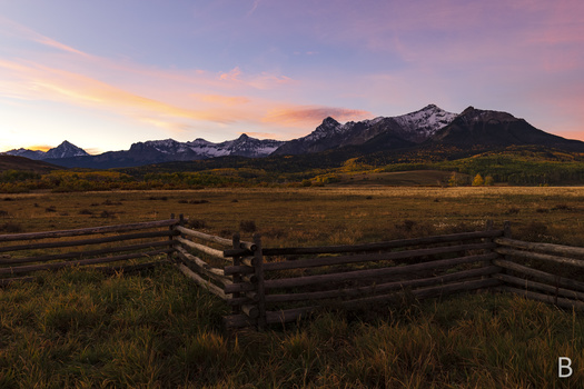 Weathered log fence in grassy meadow with snow-capped mountains at sunset.