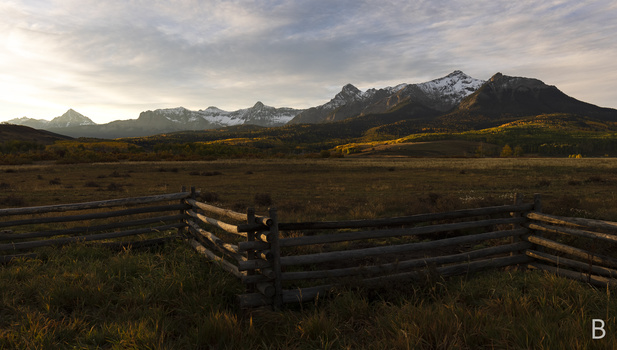 Weathered wooden fence in foreground with snow-capped mountains and golden-hour meadow in background.