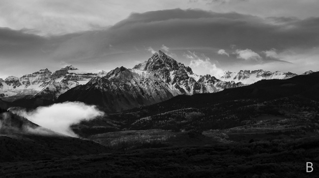 Snow-capped mountain peaks rising above forested slopes with low-lying clouds and dramatic sky.