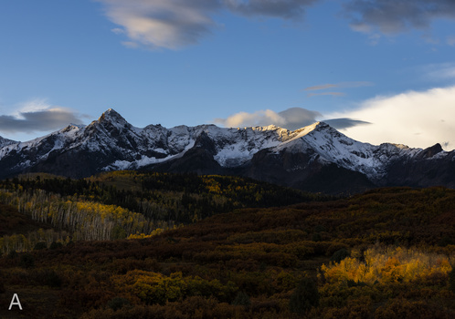 Snow-capped mountain peaks with golden light at sunset, surrounded by forested foothills and autumn vegetation.