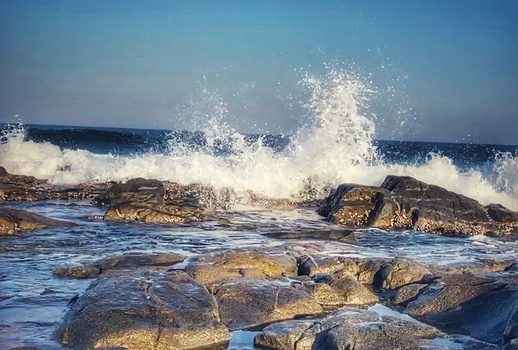 Large ocean waves crash against rocky outcrops with spray and foam in bright daylight.