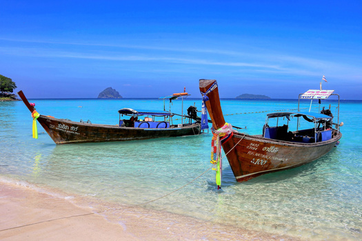 Two wooden longtail boats anchored in shallow turquoise water off a white sand beach.