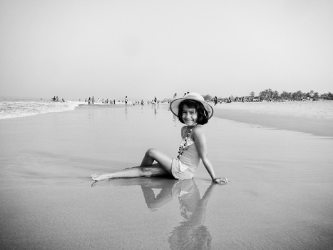 Woman in wide-brimmed hat sitting in shallow ocean water, wearing light clothing and posing for camera.