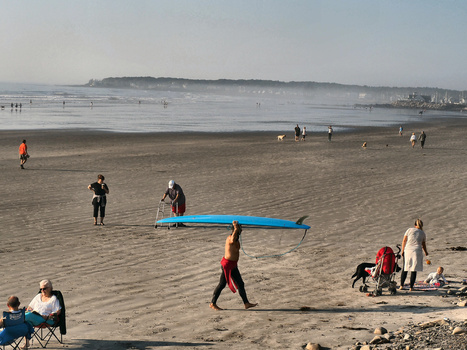 Beachgoers with blue kayak on sandy beach on overcast day with calm water and distant shoreline.