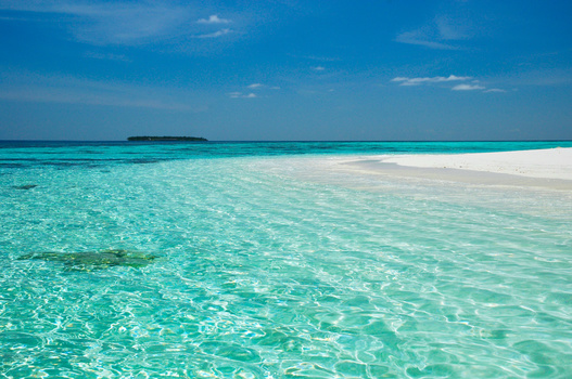 Shallow turquoise lagoon with white sand bar and distant island under clear blue sky.