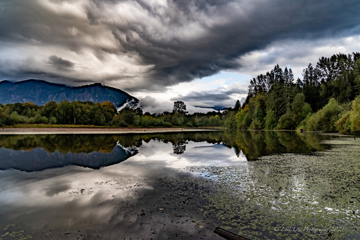 Dramatic storm clouds gather over a river valley with forested banks and distant mountains.