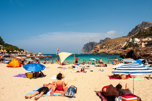 Busy Mediterranean beach with colorful umbrellas, beachgoers, and boats anchored in turquoise water beneath rocky cliffs.