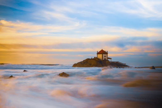 Golden hour seascape with a solitary temple structure on a rocky island surrounded by soft, blurred waves.