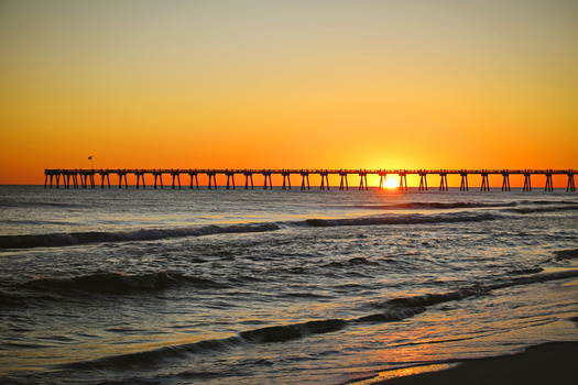 Sun setting directly beneath a long pier extending into calm ocean waters at golden hour.