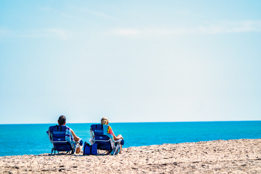 Two people sitting in beach chairs on sand facing calm turquoise water under clear sky.