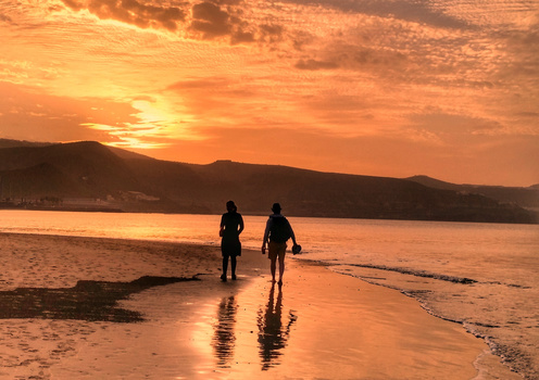 Two surfers in wetsuits stand in shallow water at sunset, facing the ocean with mountains silhouetted in the background.