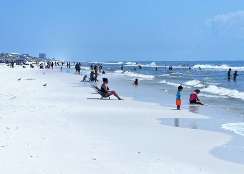 Beachgoers swimming and wading in ocean waves on a sunny day with white sand beach and clear blue sky.