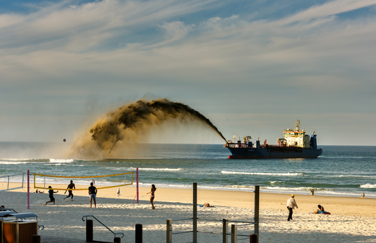 Large cargo ship offshore with massive sand plume erupting from its hull during controlled beaching operation.