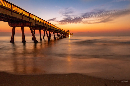 Fishing pier extending into the ocean during golden hour sunset with warm lighting on the structure.