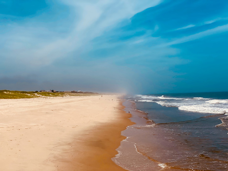 Wide sandy beach with gentle waves under a dramatic blue sky with wispy clouds.