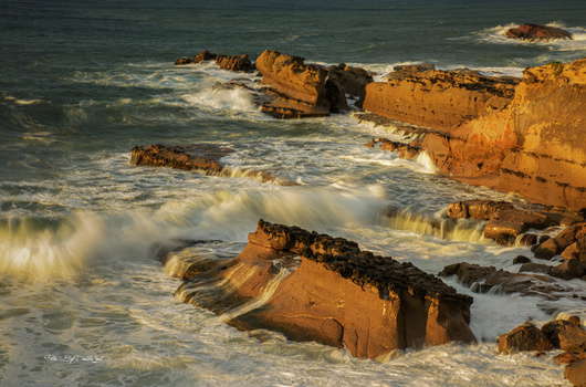 Waves crash against golden rocky outcrops at sunset, creating white foam in a dramatic coastal seascape.