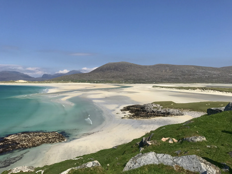 Turquoise lagoon with white sand beach backed by green meadow and distant mountains under clear blue sky.