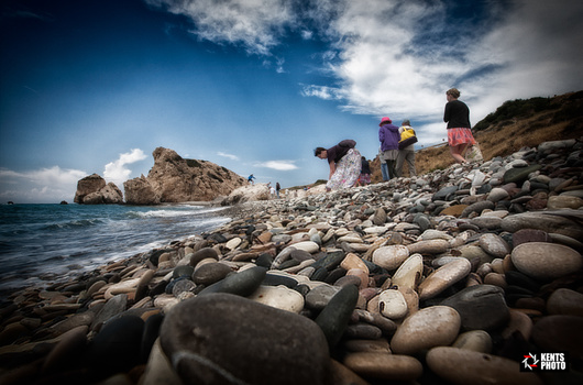 Tourists exploring a rocky beach with a large rock formation visible offshore under a blue sky with white clouds.