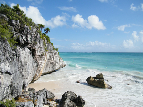 Limestone cliff with tropical vegetation overlooking turquoise ocean and white sand beach.