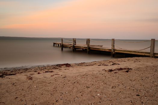 Wooden pier extending into calm water at sunset with sandy beach in foreground.