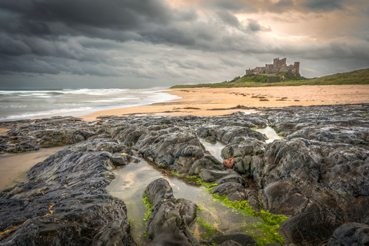 Rocky tidal pools with green algae in foreground, sandy beach and castle ruins under dramatic cloudy sky.