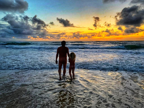 Adult and child standing in shallow ocean water at sunset with dramatic golden and orange sky.