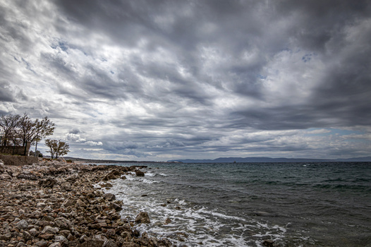Rocky shoreline with bare trees under dramatic storm clouds and choppy water.