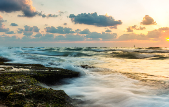 Waves crash against moss-covered rocks at sunrise, with golden light breaking through clouds over the ocean.