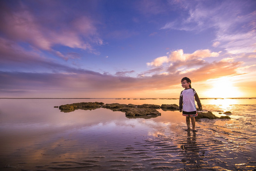 Child standing in shallow water at sunset with rocky outcrops visible offshore.