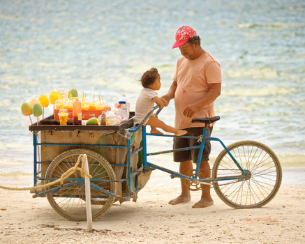 Man and child at a beach fruit cart on a bicycle with golden hour lighting.
