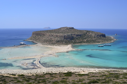 Aerial view of a turquoise lagoon with a rocky island, sandy beaches, and anchored boats under clear sky.