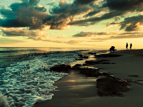 Rocky coastline at golden hour with dramatic storm clouds and silhouetted figures on the beach.