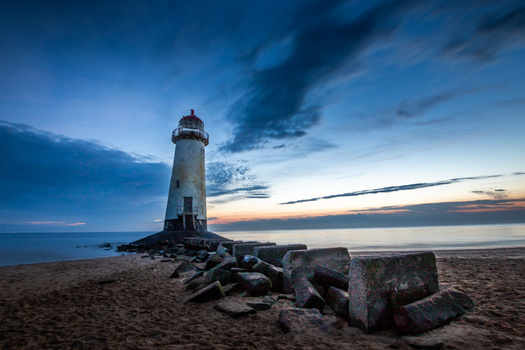 Lighthouse standing on rocky breakwater at dusk with dramatic blue sky and calm water.